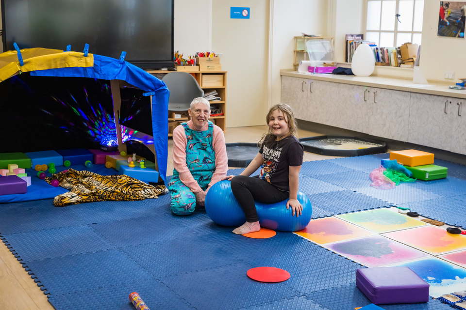 Indoor sensory playroom with colourful mats, foam blocks, and a tent-like structure with lights. A child sits on a blue exercise ball next to an adult, engaging in sensory and physical activities.